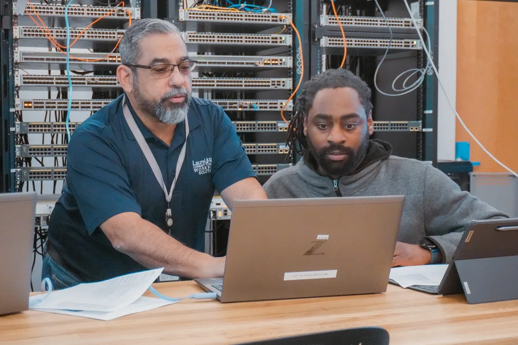 An instructor and student working at a laptop in front of networking equipment racks