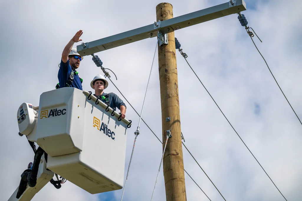 Two power line trainees in a bucket truck beside a utility pole while an instructor gestures upward